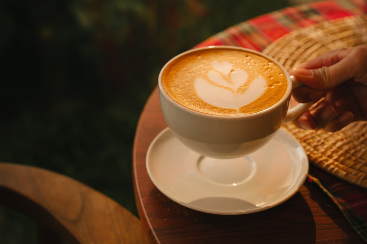 Latte art coffee cup on wooden table with hand holding cup, warm cozy atmosphere with foam heart design on top