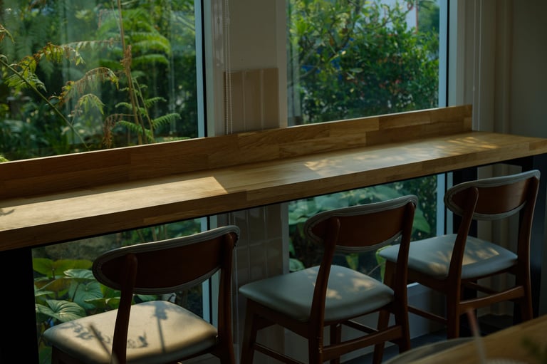 A wooden bar counter with chairs by a large window, overlooking a lush green garden in soft morning sunlight